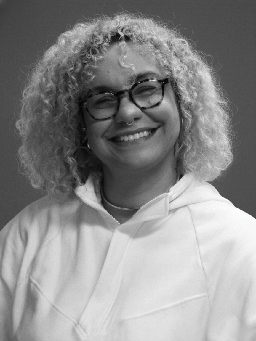 Mulher negra de pela clara, cabelos loiros e cacheados. Usa óculos está sorrindo.