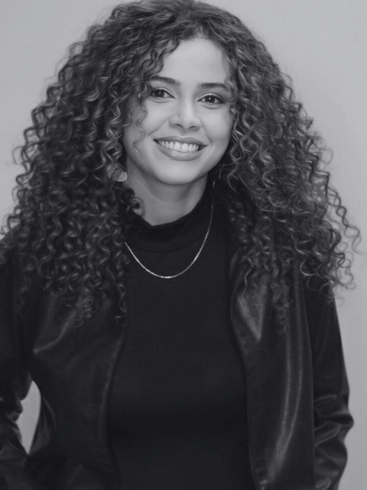 Mulher branca de cabelo cacheado abaixo do ombro está sorrindo.